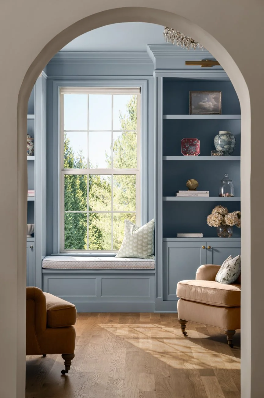 A cozy sitting area with a blue built-in window seat and bookshelf, featuring a large window with greenery outside, and two beige armchairs on a wooden floor, decorated with pillows and a chandelier above.