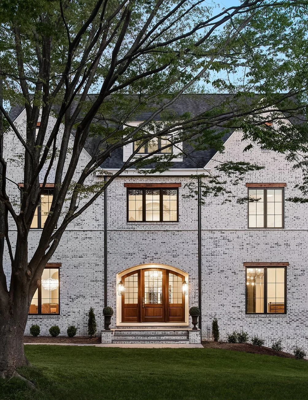 Front view of a large brick house with multiple windows, a wooden front door with glass panes, two potted plants on steps, and trees in front, illuminated by exterior lighting at dusk.