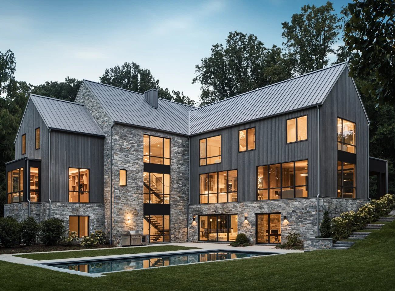 Modern multi-story house with stone and dark vertical siding exterior, large windows, and a pool in the backyard at dusk.
