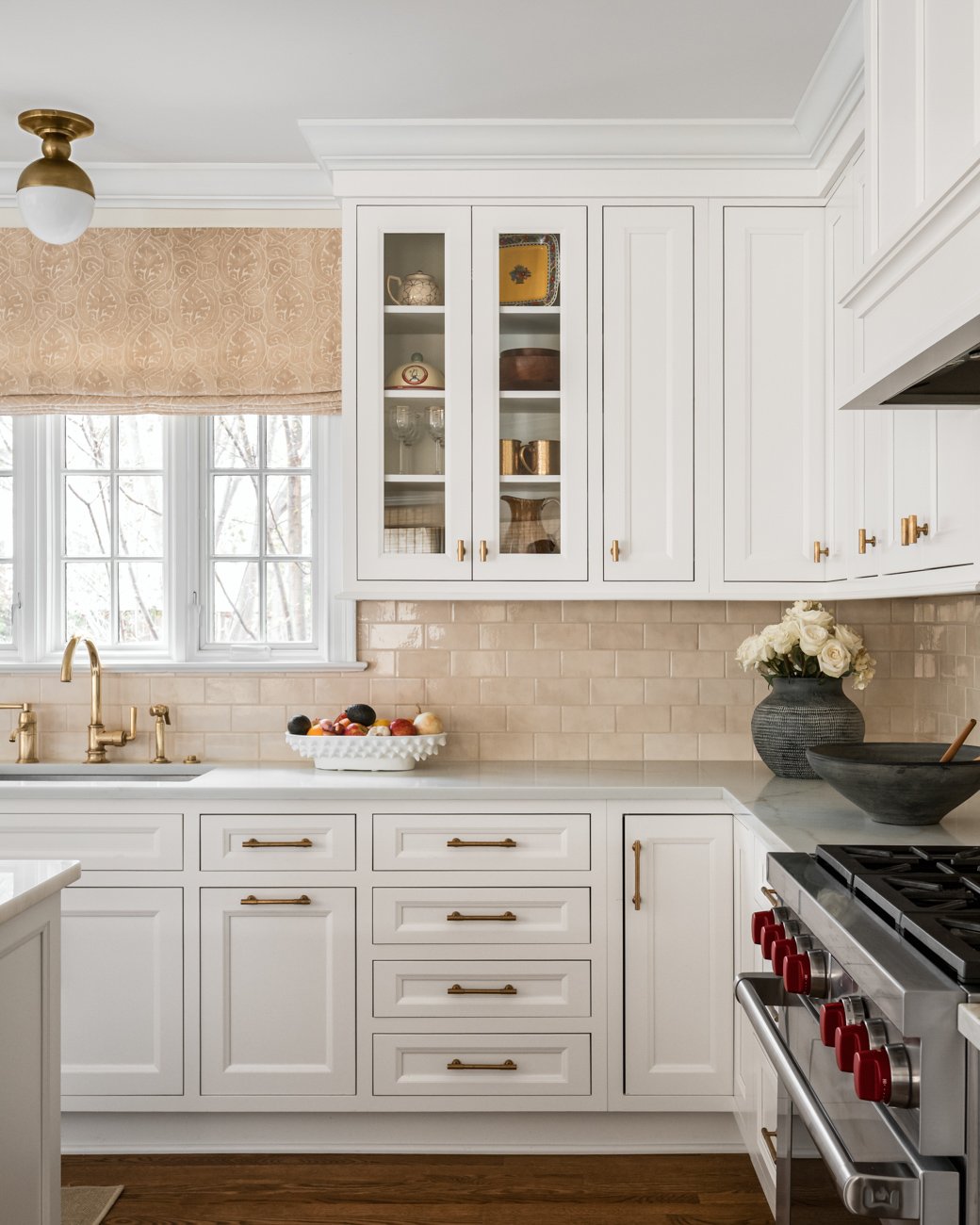 White kitchen cabinets with gold hardware, a window with beige patterned valance, countertop with a fruit bowl, black vase with white roses, black bowl with a wooden spoon, and a stove with red knobs.