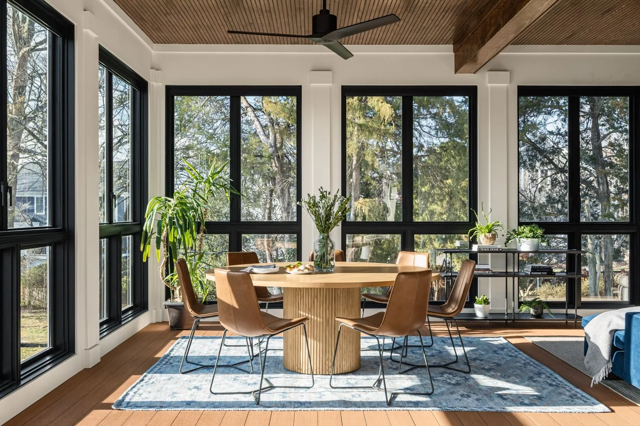 A bright dining room with large windows, a round wooden table with six brown chairs, a vase with green plants on the table, a blue patterned rug, and leafy indoor plants near the windows.