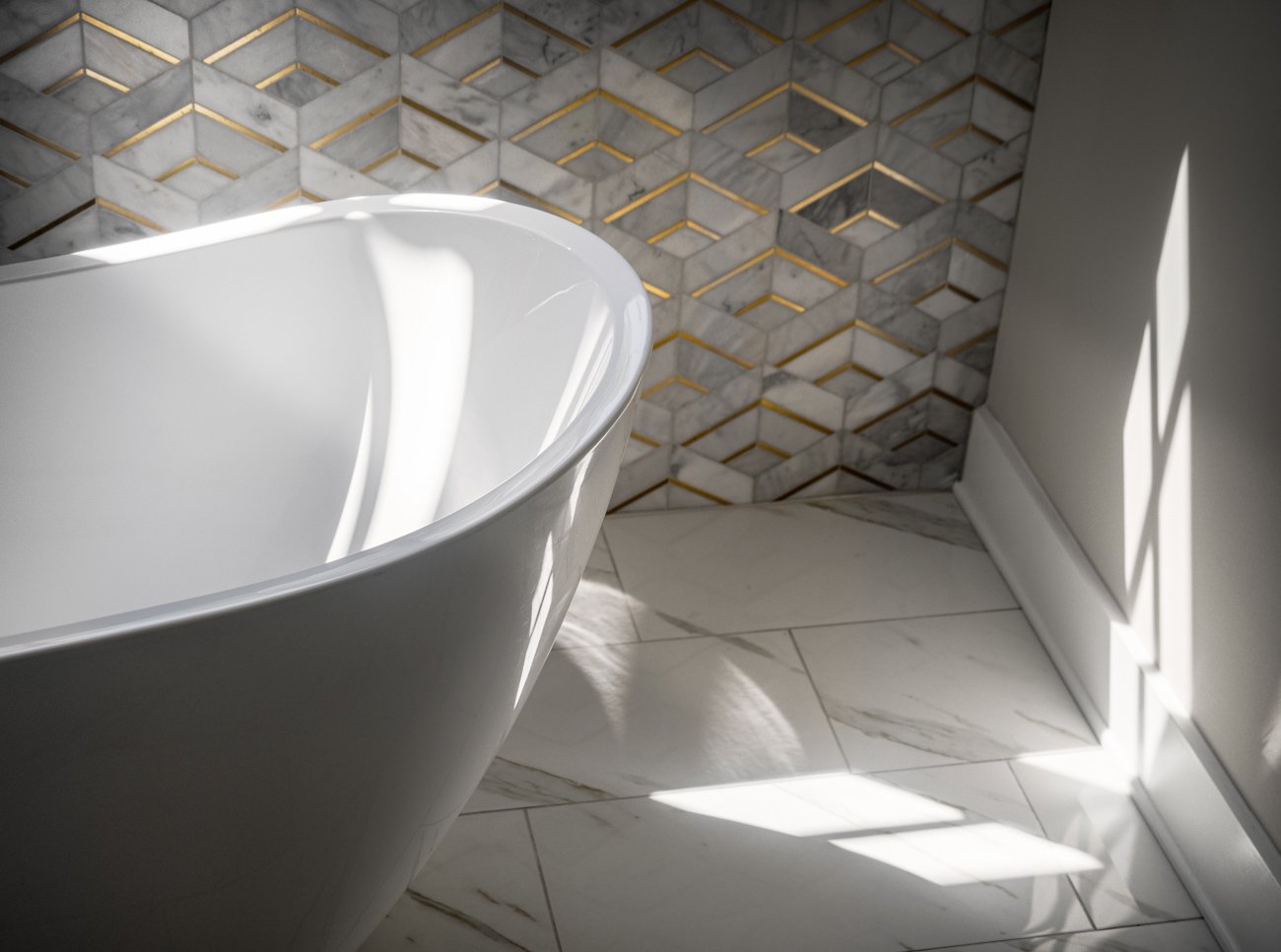 Close-up of a white freestanding bathtub in a bathroom, with sunlight casting shadows on the marble-tiled floor and wall.