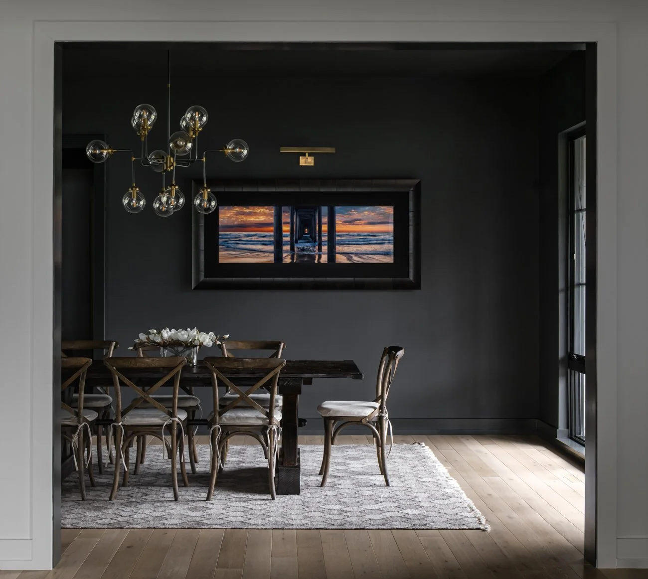 Dining room with wooden table and chairs, dark walls, chandelier, ocean sunset picture on the wall, and windows on the side.