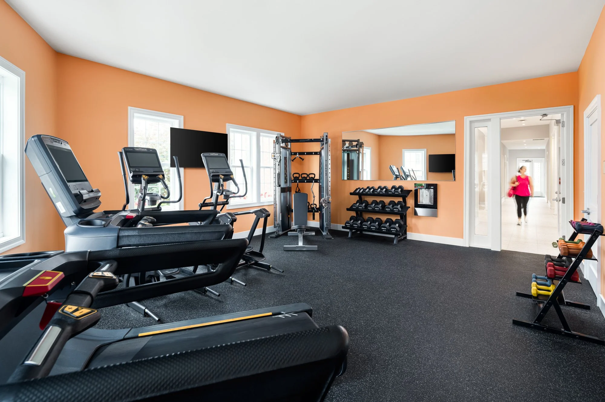 Empty fitness room with exercise machines, free weights, and an orange wall, with a person walking in the background hallway.
