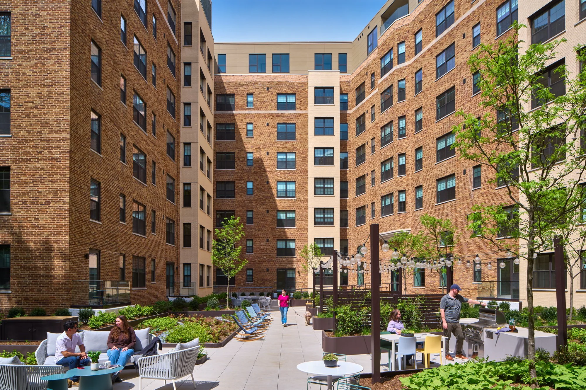Outdoor courtyard with people relaxing and socializing, surrounded by tall brick apartment buildings. There are trees, plants, and outdoor furniture, with some people sitting on chairs and one person playing a barbecue grill.