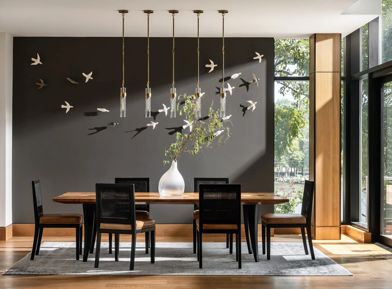 Modern dining room with a wooden table, six black chairs, a white vase with green branches, a black accent wall with bird sculptures, and large windows letting in natural light.