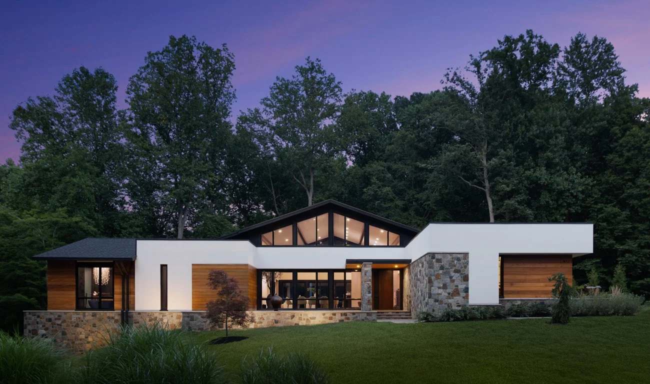 Modern house with large glass windows, stone and wood exterior, situated in a lush green yard with trees in the background at dusk.