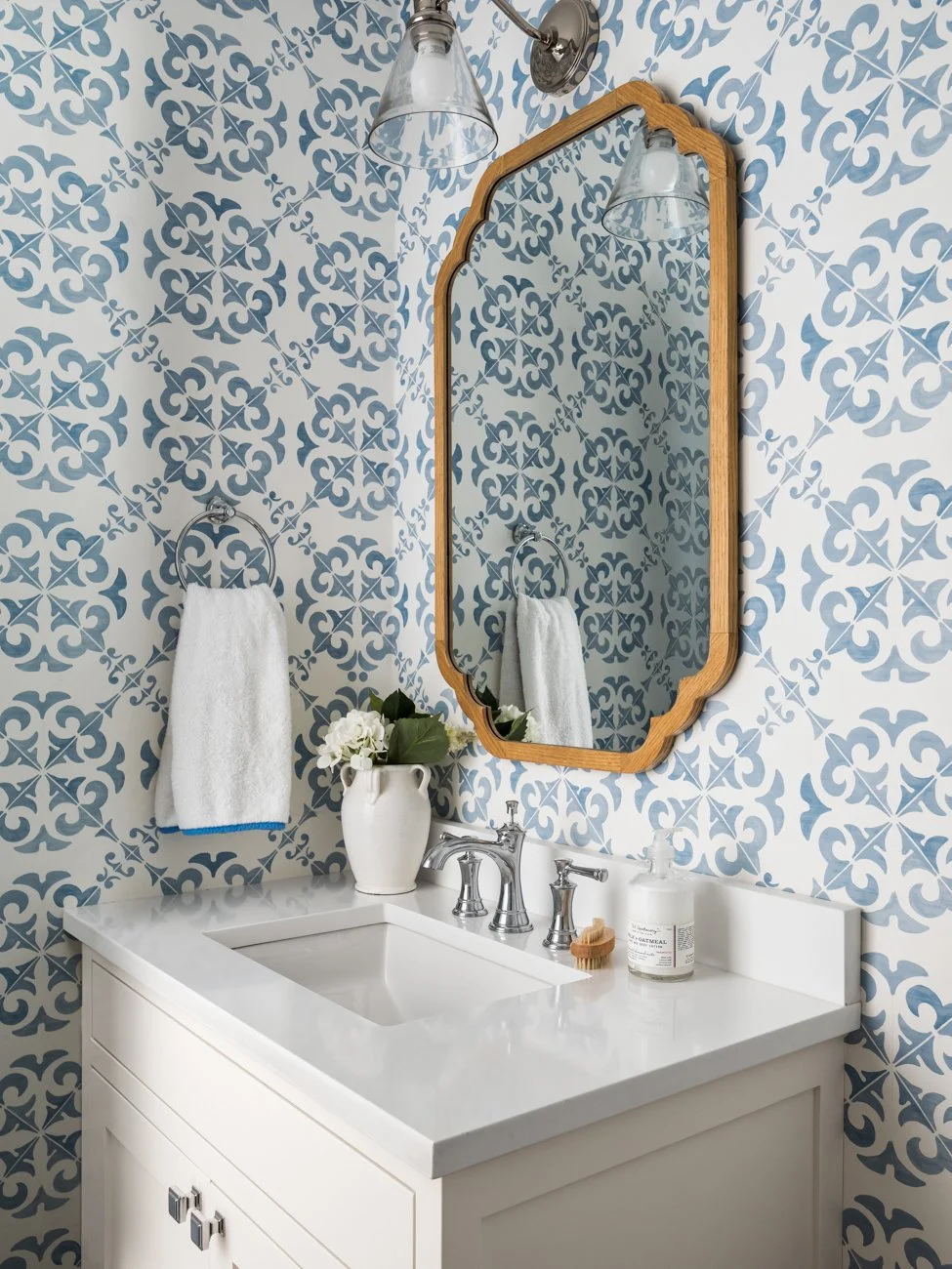 Bathroom vanity with a white countertop, a white towel on a circular silver towel ring, a ceramic vase with white flowers and green leaves, a soap dispenser, and a wooden-framed mirror. The wall behind is patterned with blue and white wallpaper.