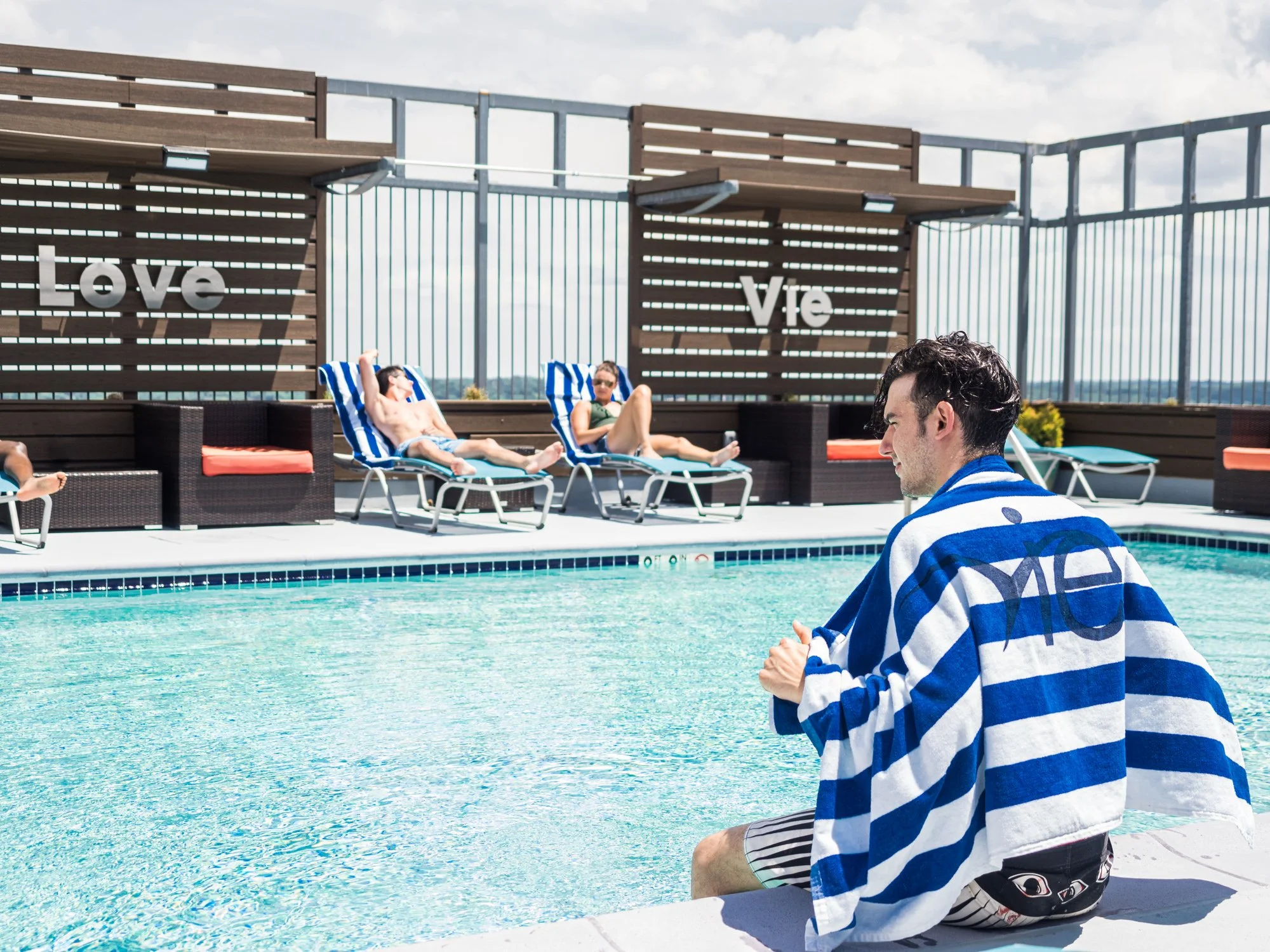 A man sitting by a rooftop swimming pool with a towel draped around his shoulders, with other people lounging on deck chairs in the background, and the words 'Love' and 'Vie' displayed on wooden wall panels.