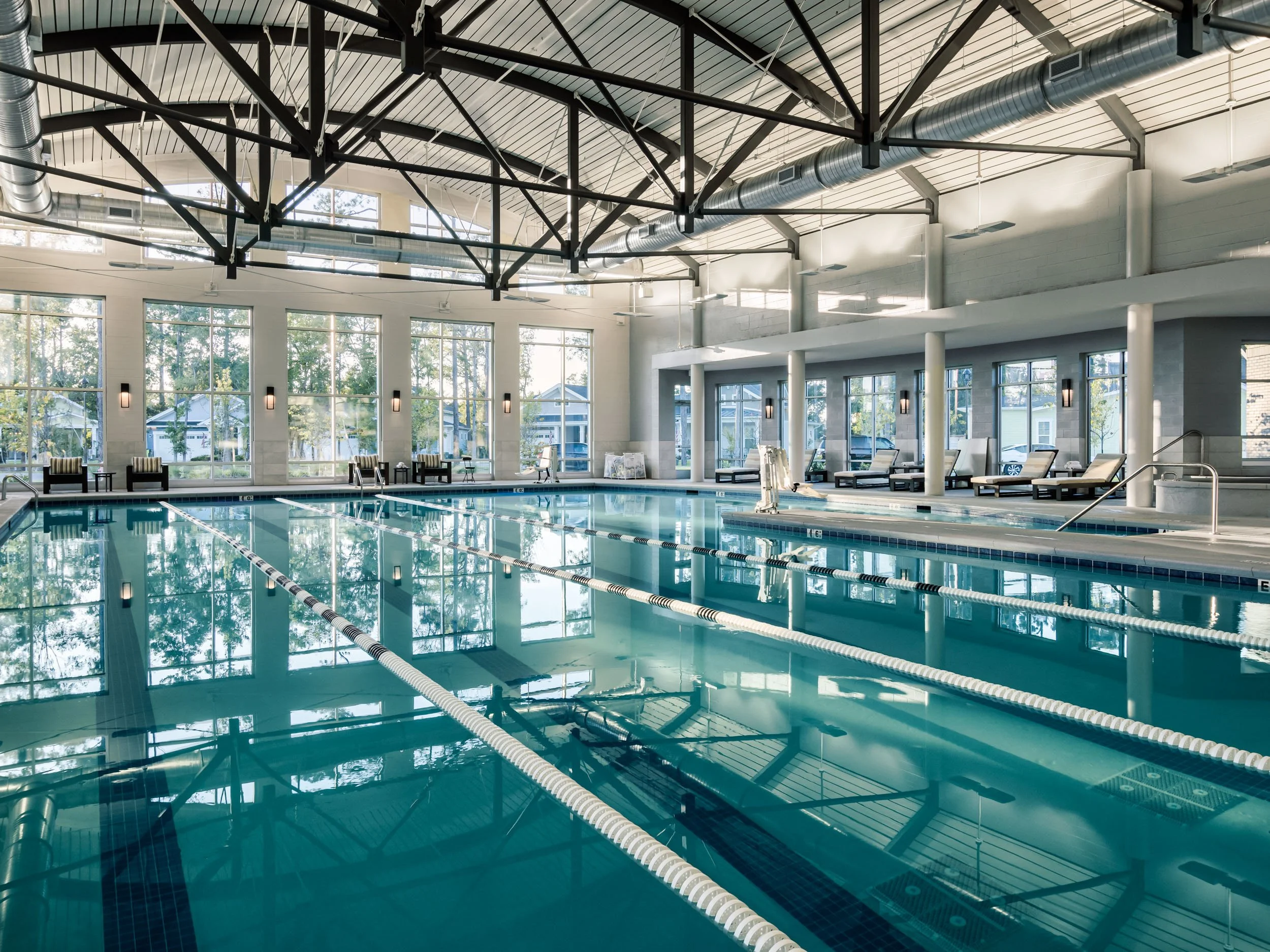 Indoor swimming pool with multiple lanes, lounge chairs, large windows, and industrial ceiling with exposed ductwork.