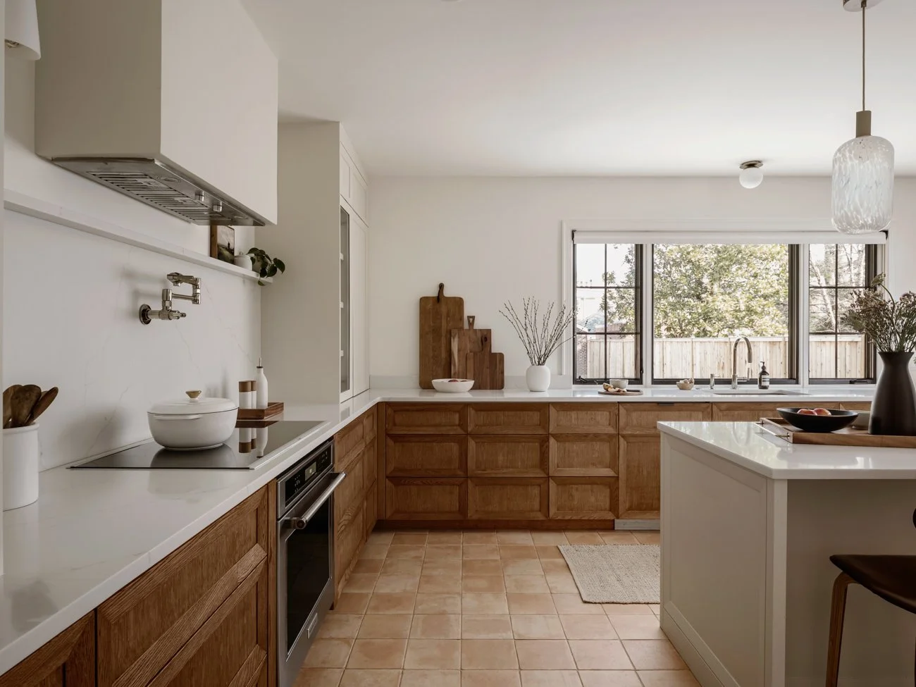 Modern kitchen with white and wooden cabinetry, large window with view of trees, beige tiled floor, kitchen island, and minimal decor including bowls, cutting boards, and vases with branches.