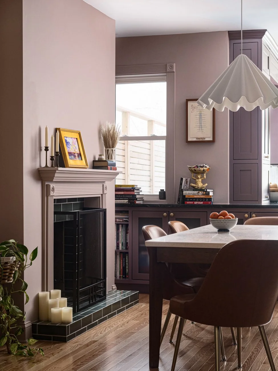 A cozy dining room with a fireplace, a window letting in natural light, and a dining table with chairs. Decor includes candles, books, and a bowl of oranges.