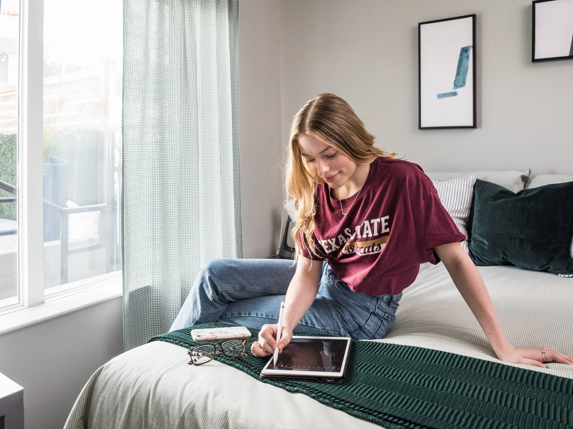 A young woman with long blond hair sitting on a bed, using a stylus on a tablet.