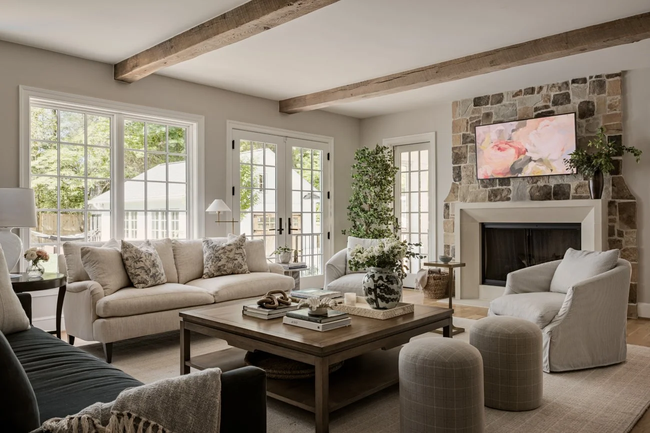 Living room with beige sofa, white armchair, stone fireplace, large windows, and wooden ceiling beams.