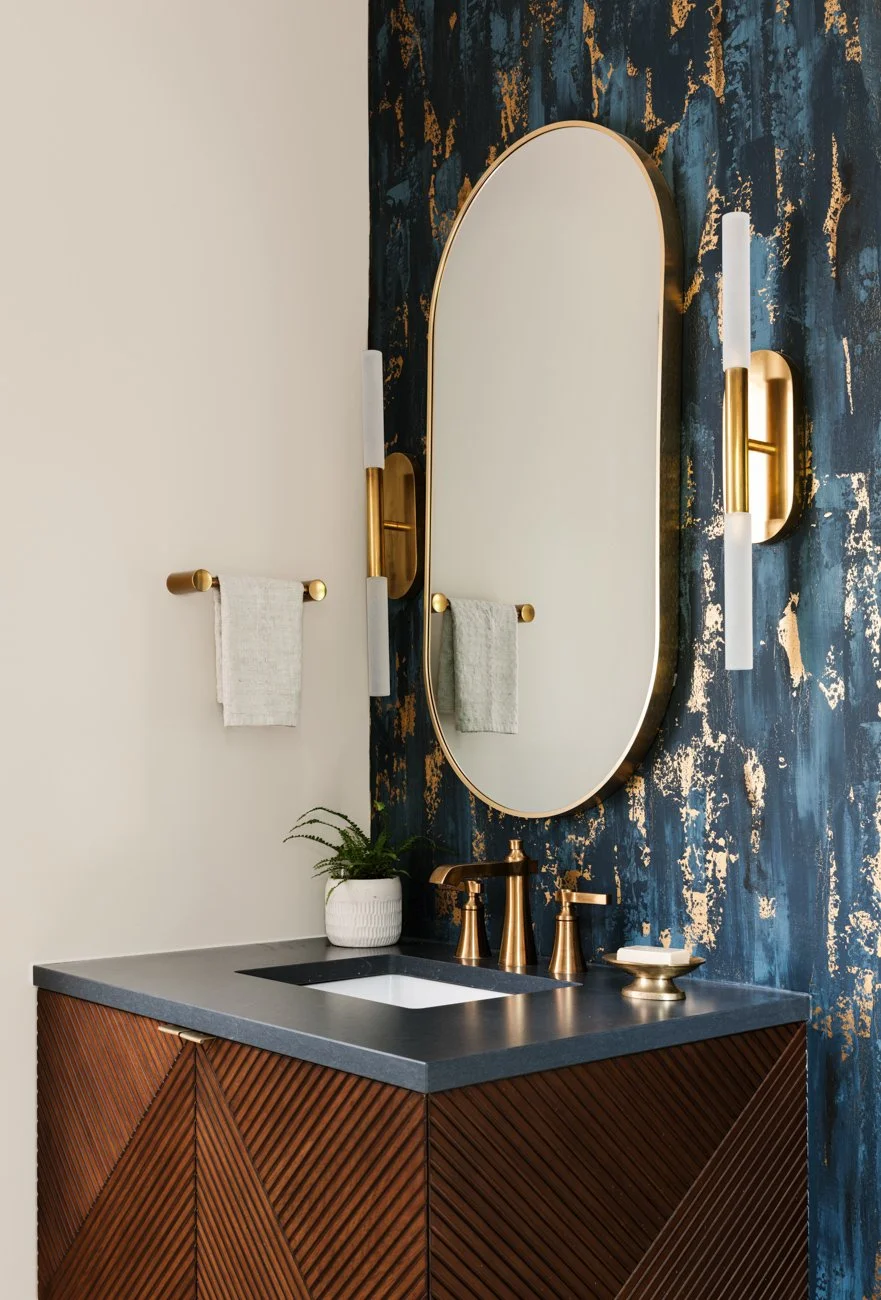 A modern bathroom vanity with a black countertop, bronze fixtures, a round mirror with a thin bronze frame, gold wall sconces, a small towel bar with a white towel, and a potted plant against a dark blue wall with gold accents.