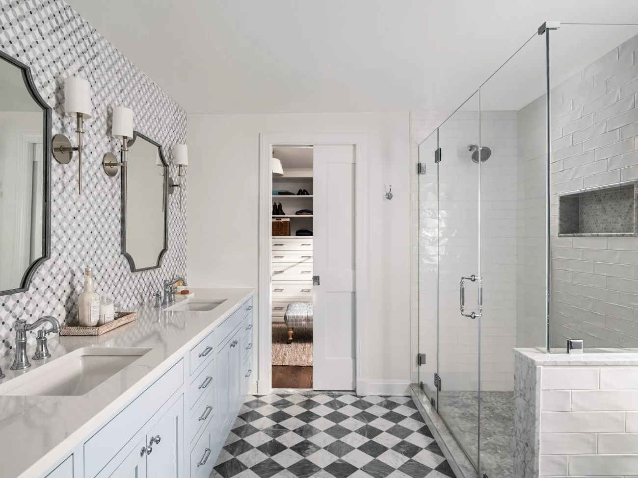 Modern bathroom with a white double vanity, patterned wall mirrors, wall sconces, and a glass-enclosed shower with white subway tiles and a built-in niche, featuring black and white checkered tile flooring.