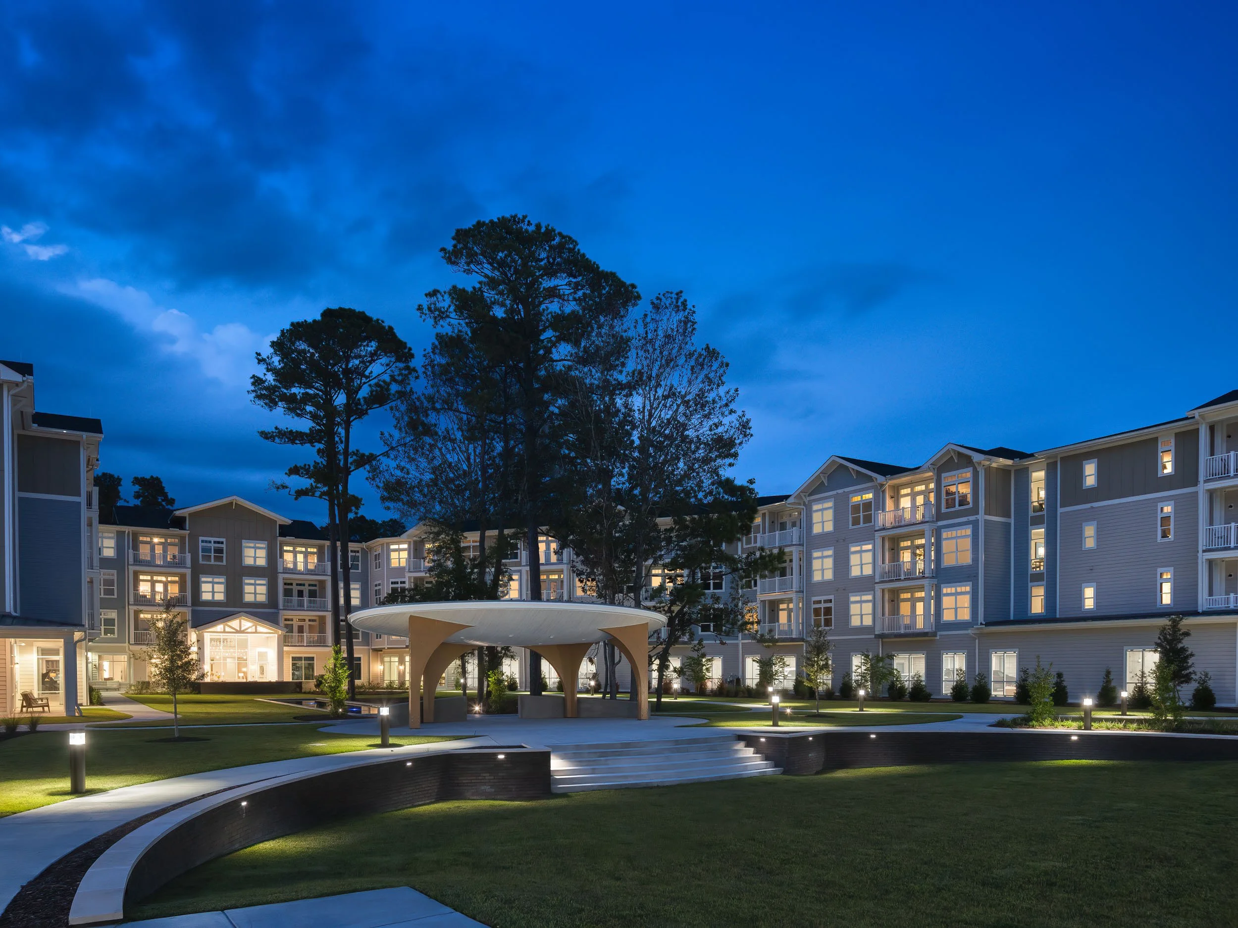 Modern apartment complex at dusk with lit windows, surrounding green lawn, trees, and a covered pavilion.