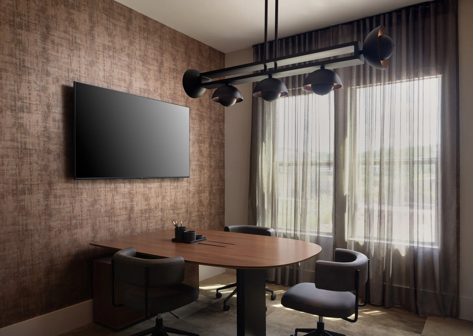 Modern office conference room with a wooden table, three black chairs, a large flat-screen TV on a textured brown wall, and natural light coming through sheer curtains on a glass window