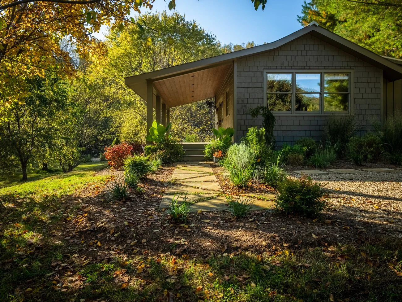 A small house with gray shingles, gravel pathway, and lush green garden surrounded by trees with yellow and orange leaves in autumn.