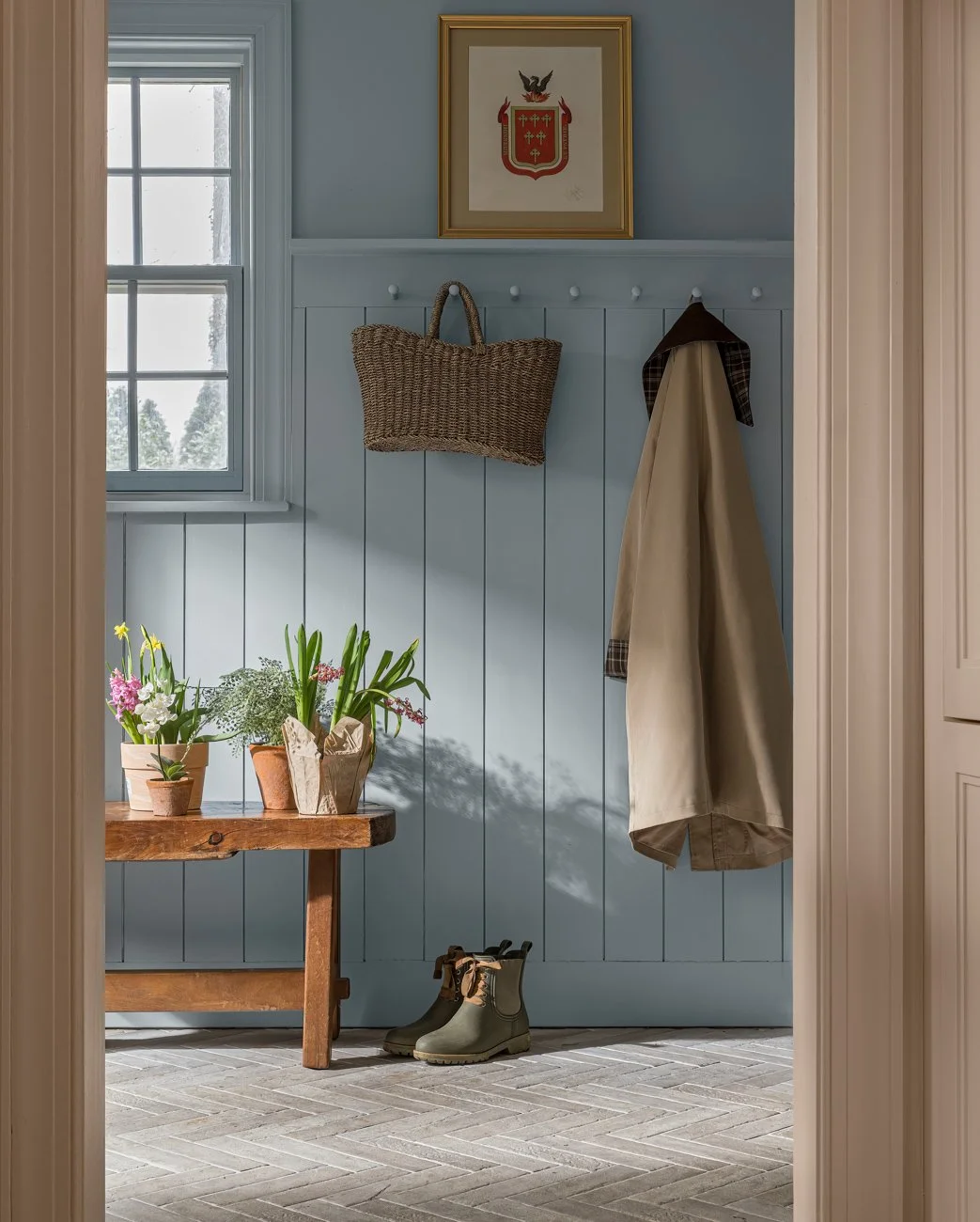 Interior view of a mudroom with a light blue wooden wall, featuring a framed crest above hooks holding a wicker bag and a coat, a window on the left, a wooden bench with potted plants, a pair of green boots underneath, and light-colored tile flooring