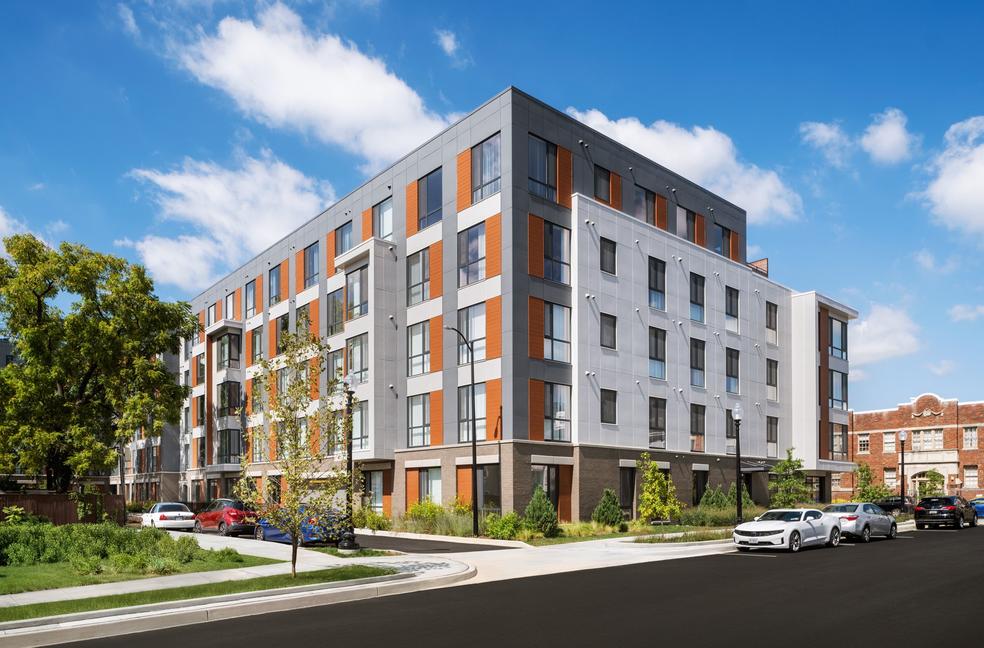 Modern multi-story apartment building with a mix of gray, white, and wood exterior, situated along a street with parked cars and landscaped greenery, under a blue sky with clouds.