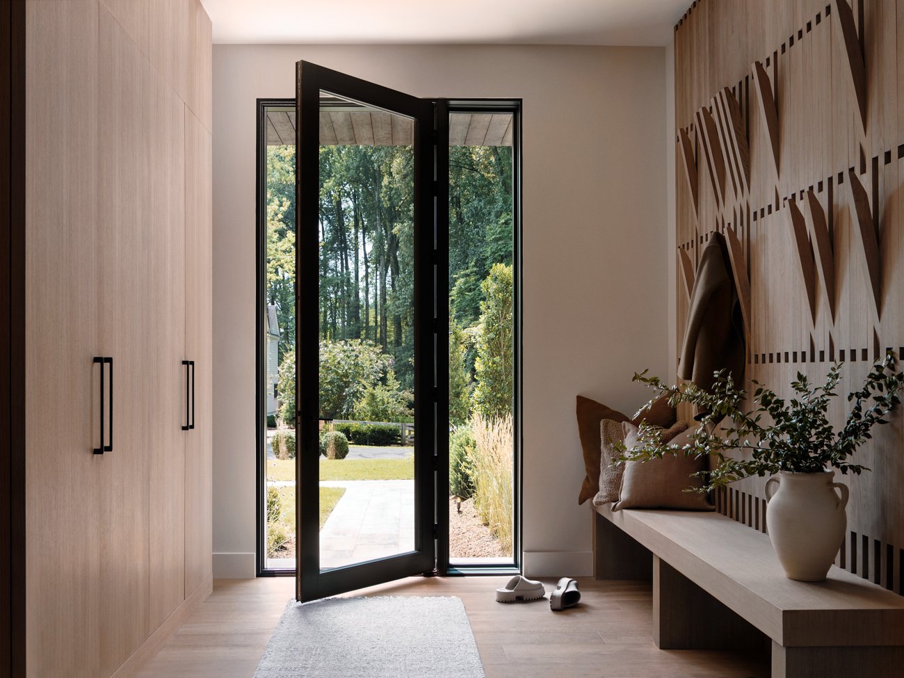 Entryway with open door to outdoor garden, wooden built-in storage, a bench with pillows, a large vase with greenery, and shoes on the floor.