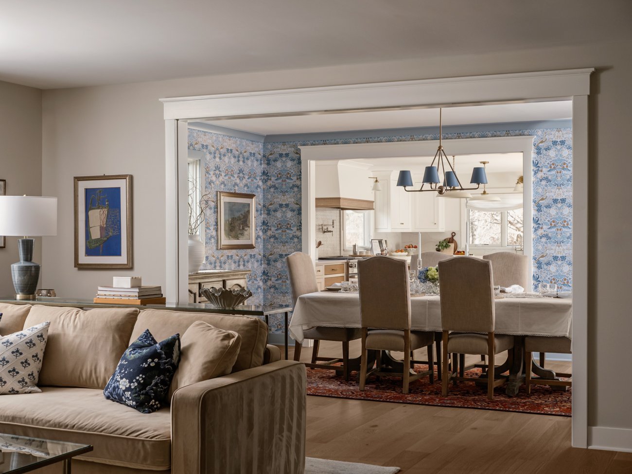 Living room with beige sofa and throw pillows, seen through doorway into dining area with six upholstered chairs around a table, blue floral wallpaper, and a chandelier over the table.