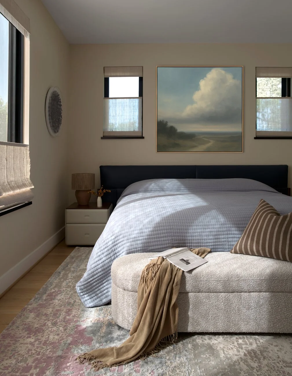 A cozy bedroom with a bed, nightstand, and decor. The bed has a dark headboard, a light-colored quilt, and a striped pillow. A blanket is draped over a beige upholstered bench at the foot of the bed. There are three small windows with shades, a frame