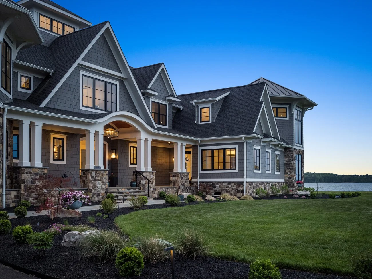 Large, elegant house with gray siding, black roofing, and a stone foundation, situated near a body of water, with a well-maintained lawn and landscaped garden.