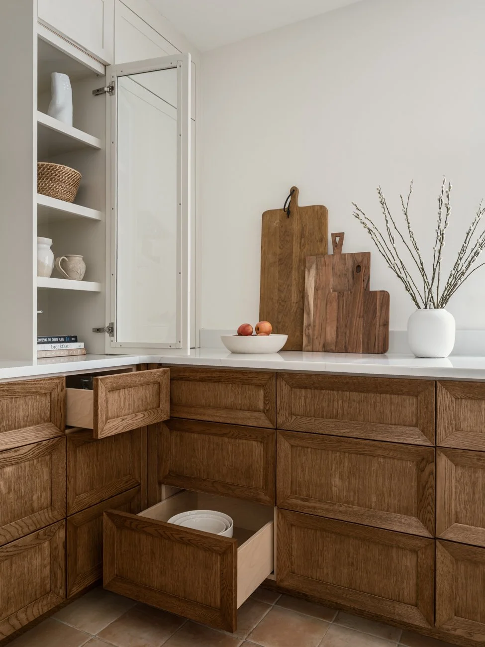 Kitchen cabinetry with open and closed drawers, cutting boards, a white bowl with apples, and a white vase with branches on a countertop.