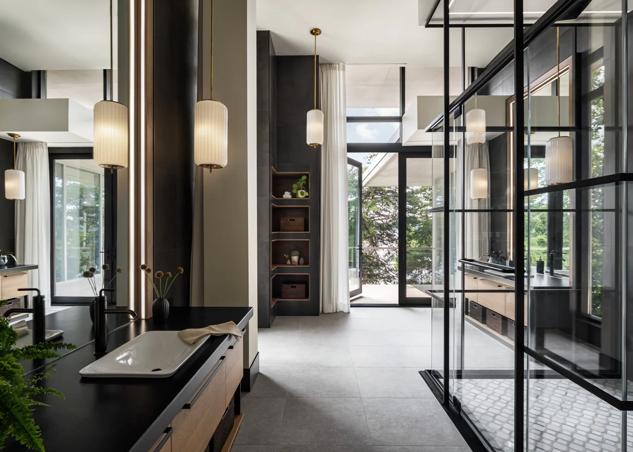 Modern bathroom with black and white accents, pendant lights, a sink with a black countertop, and a glass shower enclosure, overlooking greenery outside.