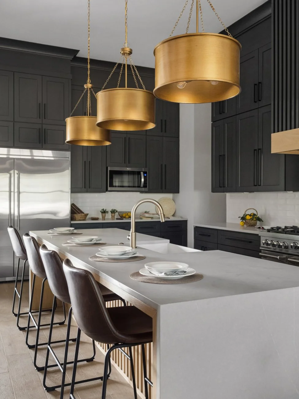 Modern kitchen with black cabinetry, stainless steel refrigerator, and white countertop island set with plates and bowls, gold hanging pendant lights, and a row of three black and brown chairs.