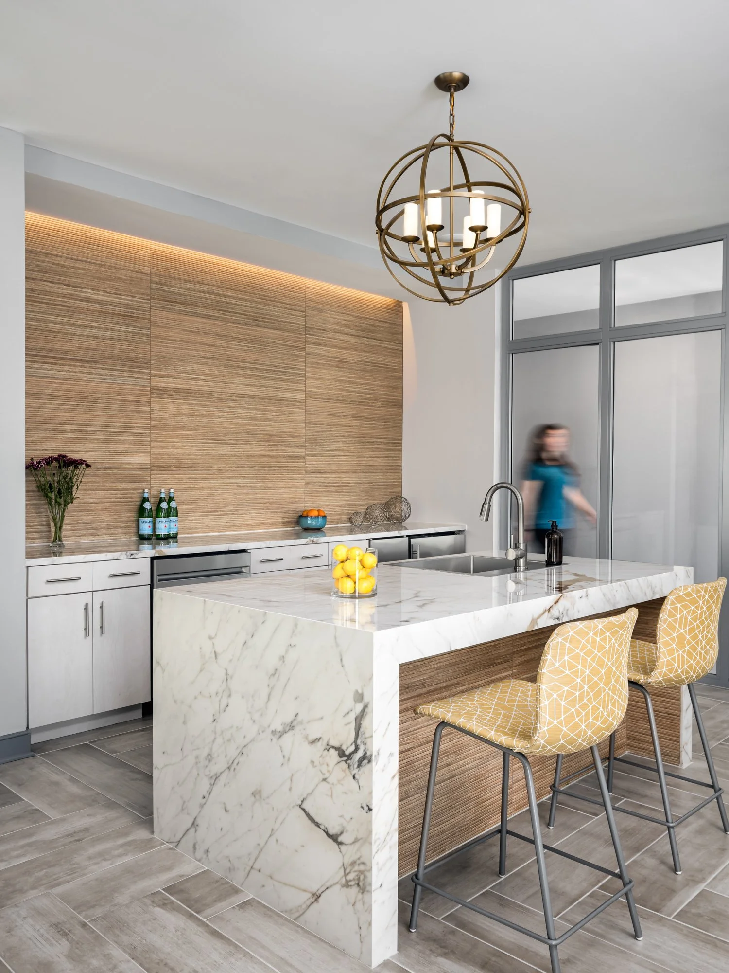Modern kitchen with a white marble island, wooden wall panel, yellow patterned chairs, and chandelier.