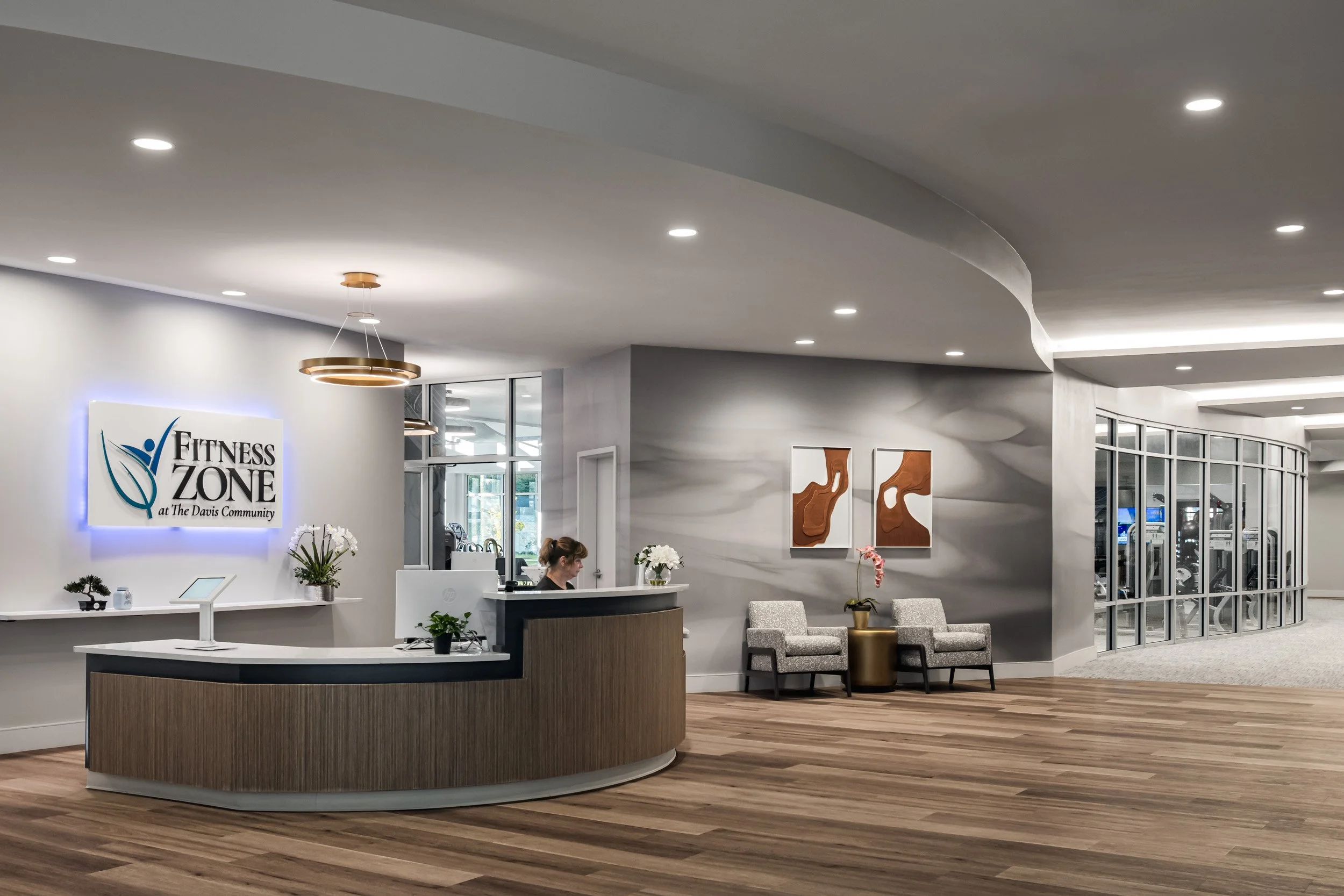 Reception area of a fitness center with a curved wooden desk, a woman working behind it, two chairs with a plant between them, wall art, and a glass wall revealing gym equipment inside.