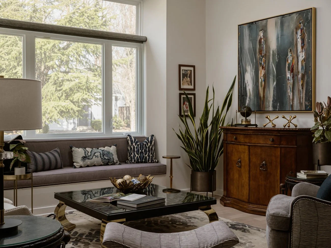 Living room with a large window, a gray sofa with decorative pillows, a black coffee table with books and a bowl, a wooden sideboard with decorative items and paintings, a tall plant, a lamp, and a patterned rug.