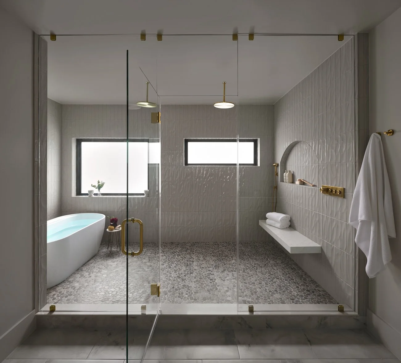 Modern bathroom with a freestanding bathtub, glass shower enclosure, and minimalistic decor, featuring stone flooring, beige tiled walls, and large windows.