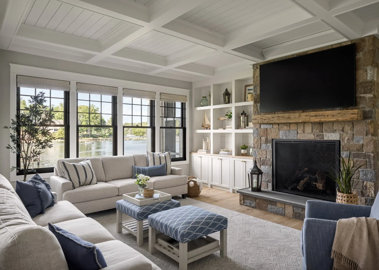 Living room with a large window, white sofas, blue and striped pillows, a stone fireplace with a mounted TV, built-in white shelves, plants, and nautical decor overlooking a lake.