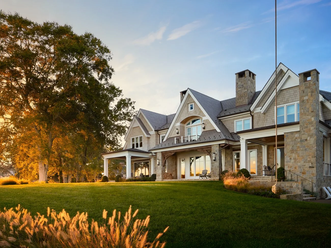 A large stone and siding house with multiple gables, large windows, and a porch, surrounded by well-maintained lawn and trees, during sunset.