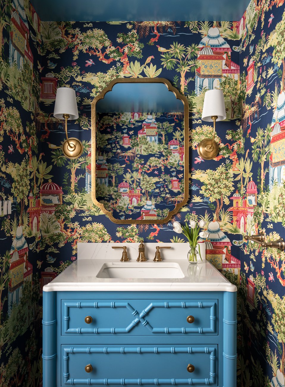 A colorful bathroom with patterned wallpaper featuring a tropical, oriental landscape, gold fixtures, a blue vanity with bamboo-style details, and a framed mirror. A vase with flowers is on the white countertop.