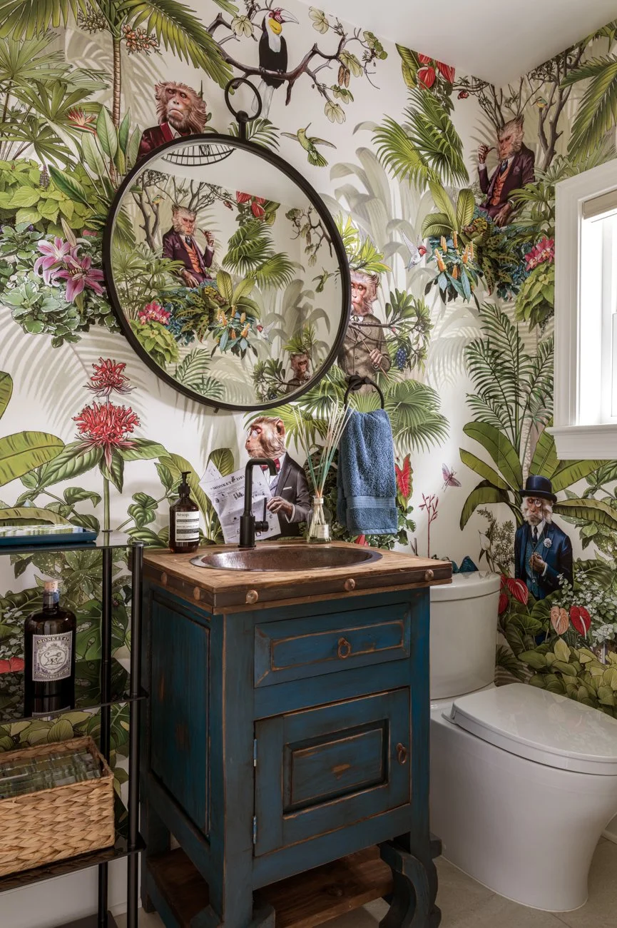 A bathroom with tropical-themed wallpaper featuring animals and people with animal heads, including lions and monkeys, dressed in vintage clothing. The bathroom contains a vintage blue vanity with a wooden countertop, a round mirror, a modern white t