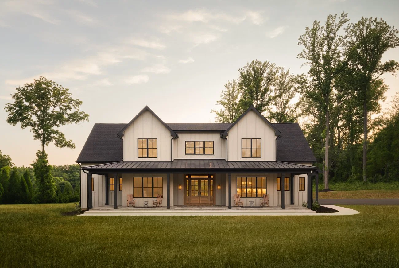Front view of a modern farmhouse style home with a large covered porch, surrounded by green grass and trees, with evening lights on.