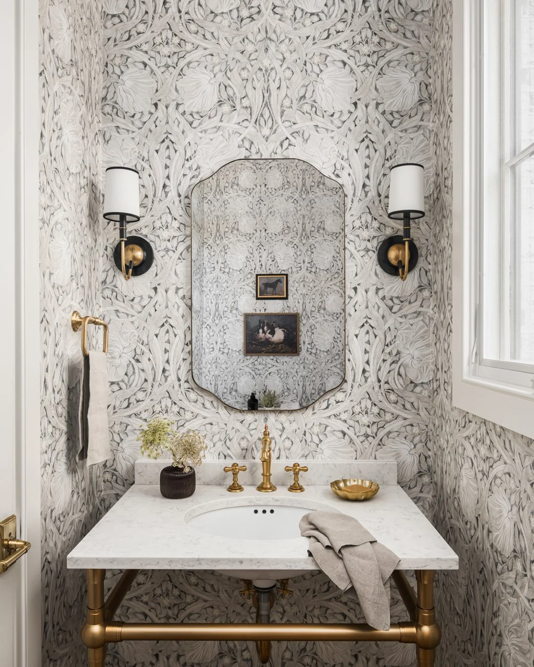 Small ornate bathroom with a white marble sink, gold fixtures, floral wallpaper, a mirror, wall sconces, and framed artwork.