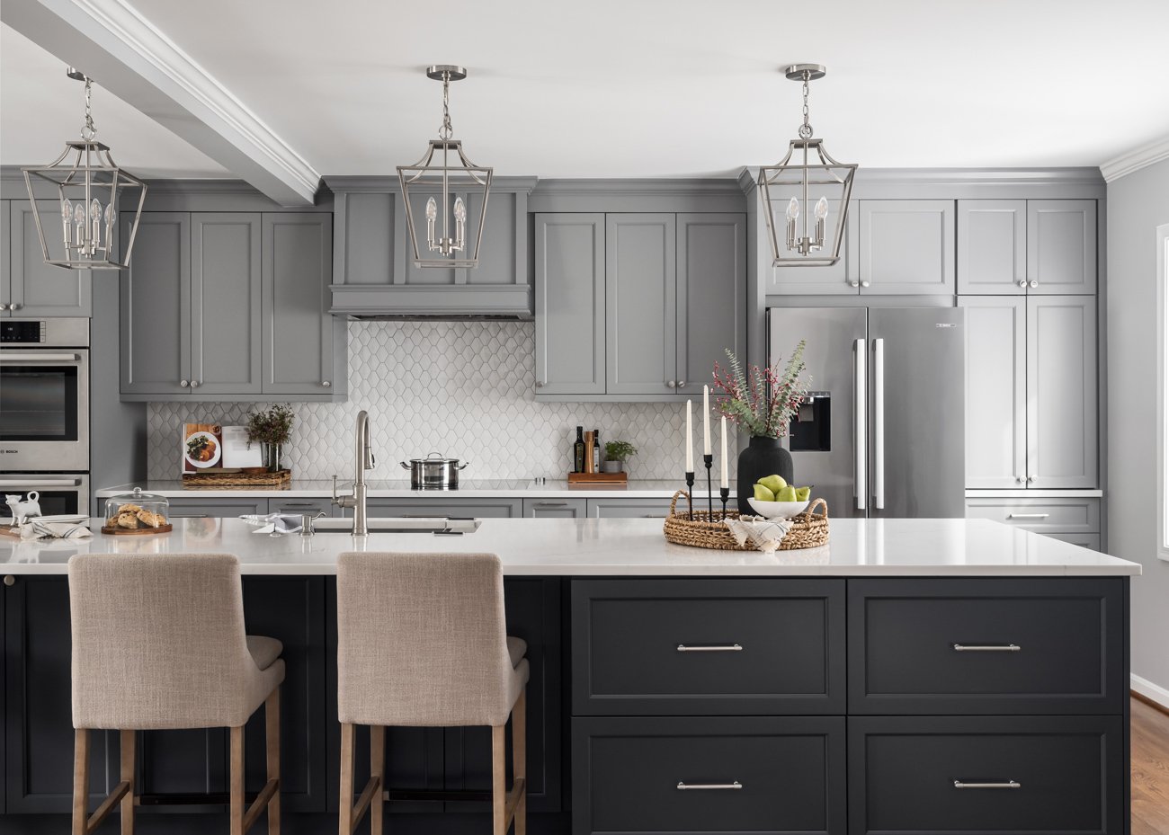 Modern kitchen with gray cabinets, white countertops, stainless steel appliances, and beige bar stools, decorated with a black vase of flowers and a bowl of green apples.
