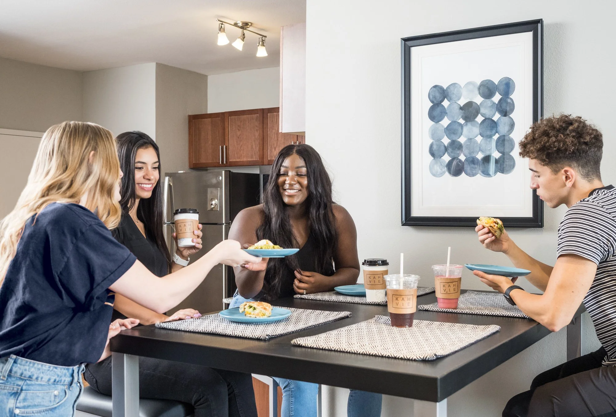 Four young adults sharing a meal and coffee at a kitchen table, smiling and chatting.