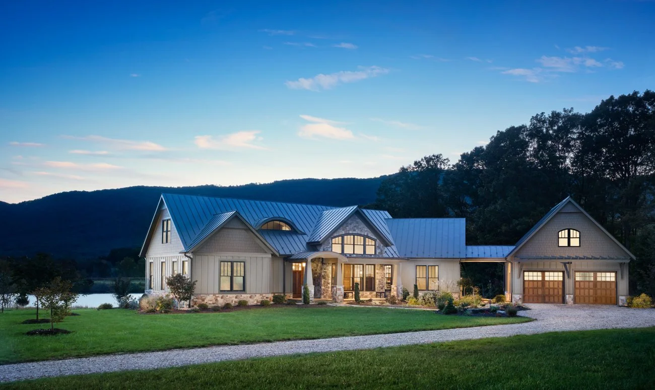 A modern house with a metal roof and large windows, surrounded by a green lawn and trees, with a mountain and river in the background during twilight.