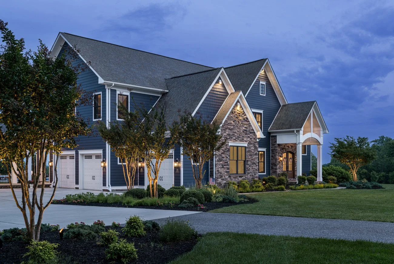A large, modern blue house with white trim, stone accents, and multiple gables, surrounded by manicured lawns and trees, during dusk with exterior lights on.