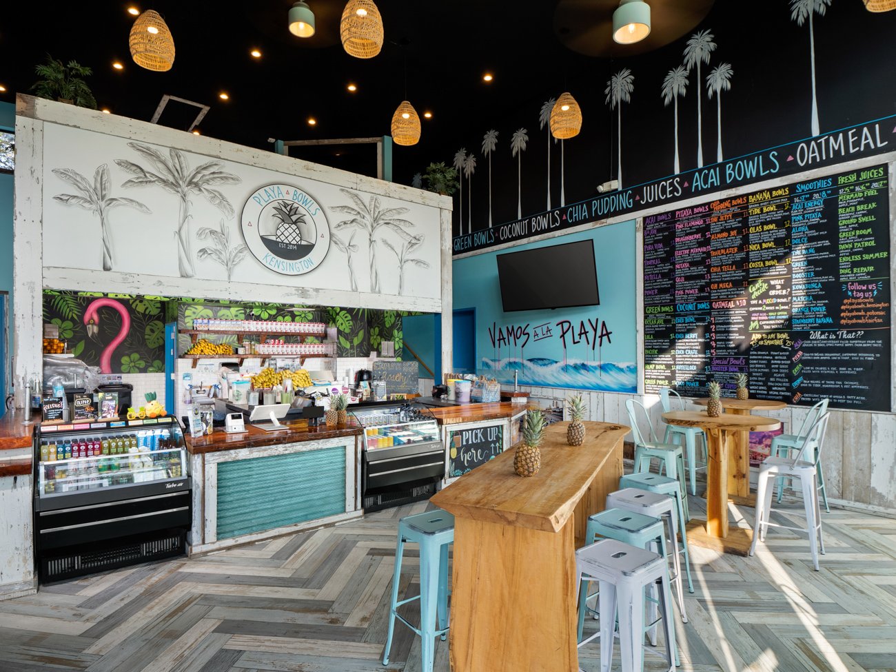 Interior of a tropical-themed cafe with a black ceiling decorated with palm tree silhouettes and hanging wicker lights. There are high tables with pineapples used as decor, a colorful menu board, and a counter with beverages and snacks. The walls are