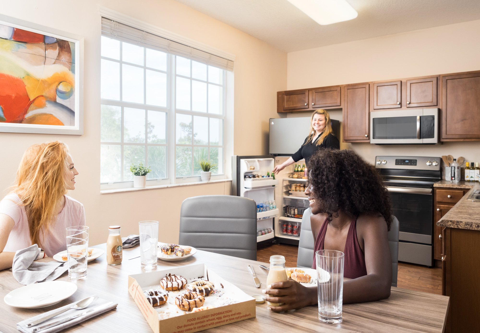 Three women sitting at a dining table with donuts and drinks, two women are smiling and talking, a woman is in the kitchen near an open refrigerator.
