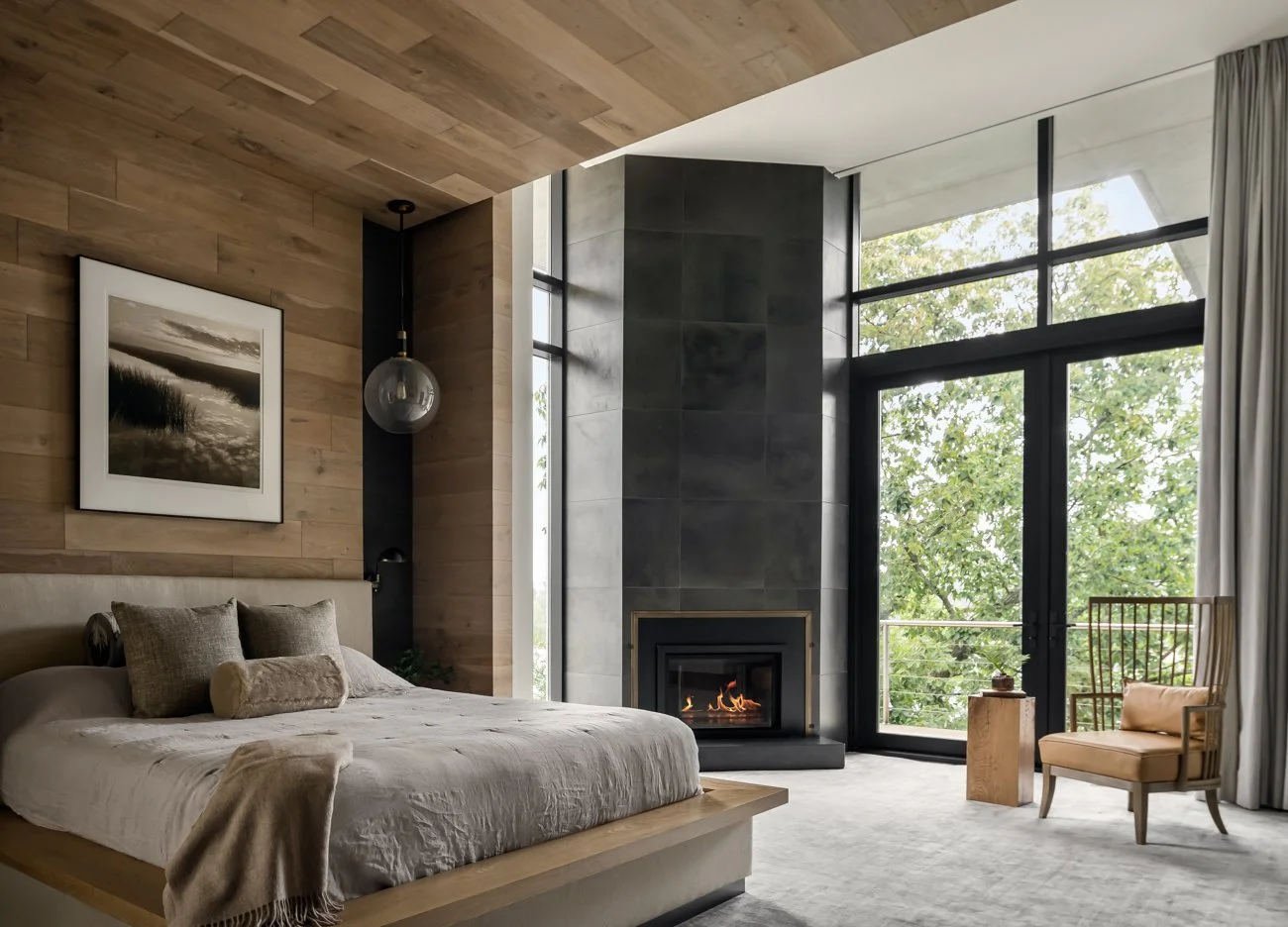 Modern bedroom with a bed, decorative pillows, a fireplace, large windows, and trees outside.