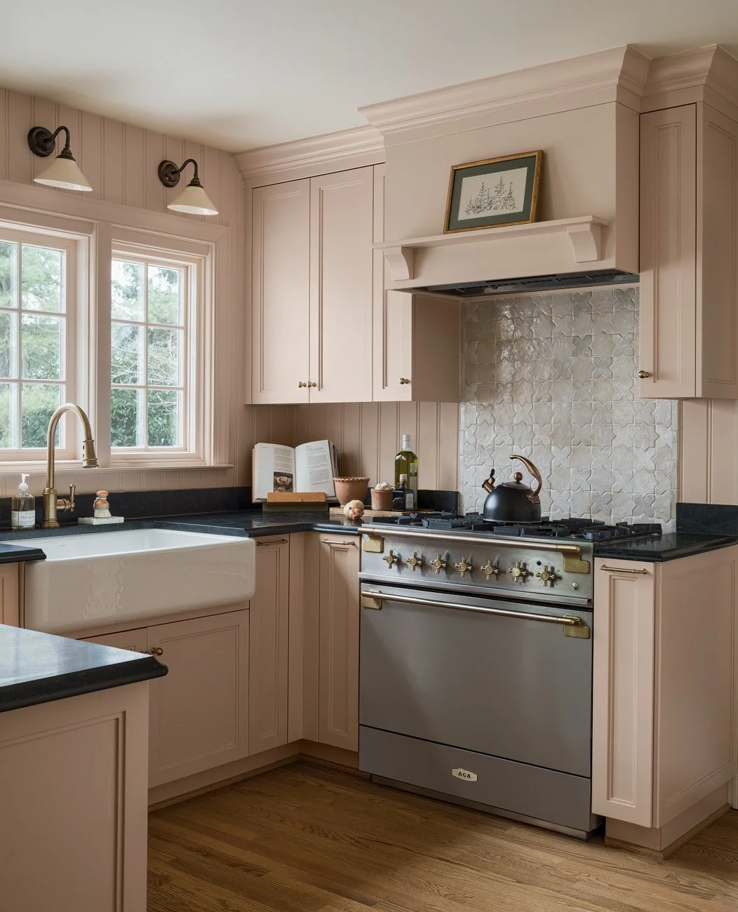 A cozy kitchen with beige cabinets, black countertops, a large farmhouse sink with a gold faucet, a window with a view of trees, a vintage stove with gold accents, a black teapot on the stove, and decorative items including an open book, bottle of ol